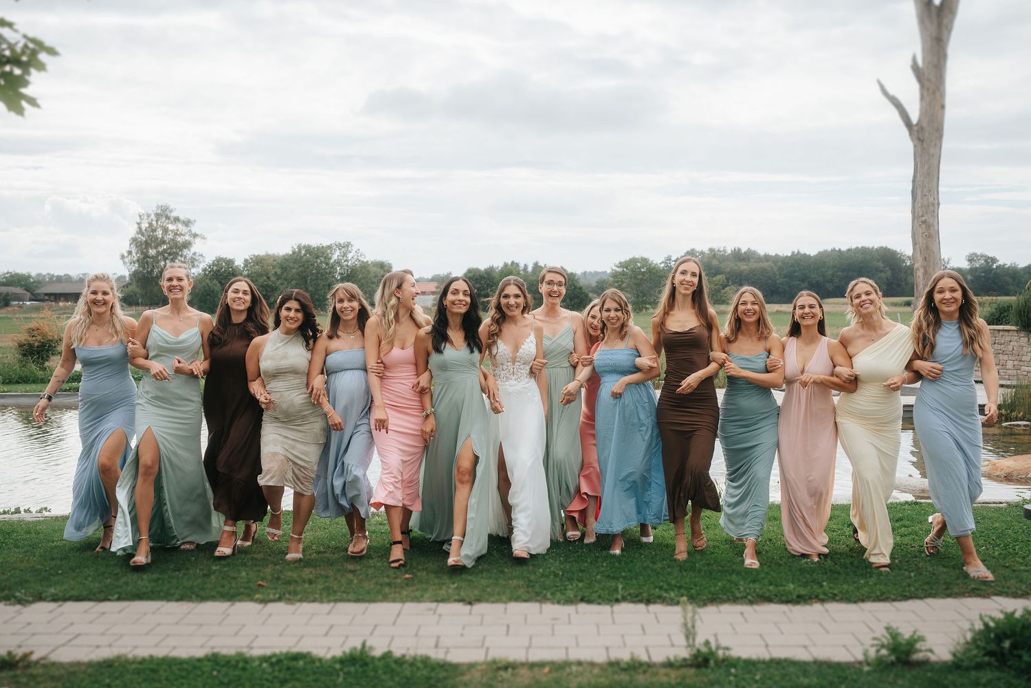 Group of women in flowing pastel and sage green dresses stand arm in arm on a grassy lawn by a lake, smiling in a bridal party portrait.