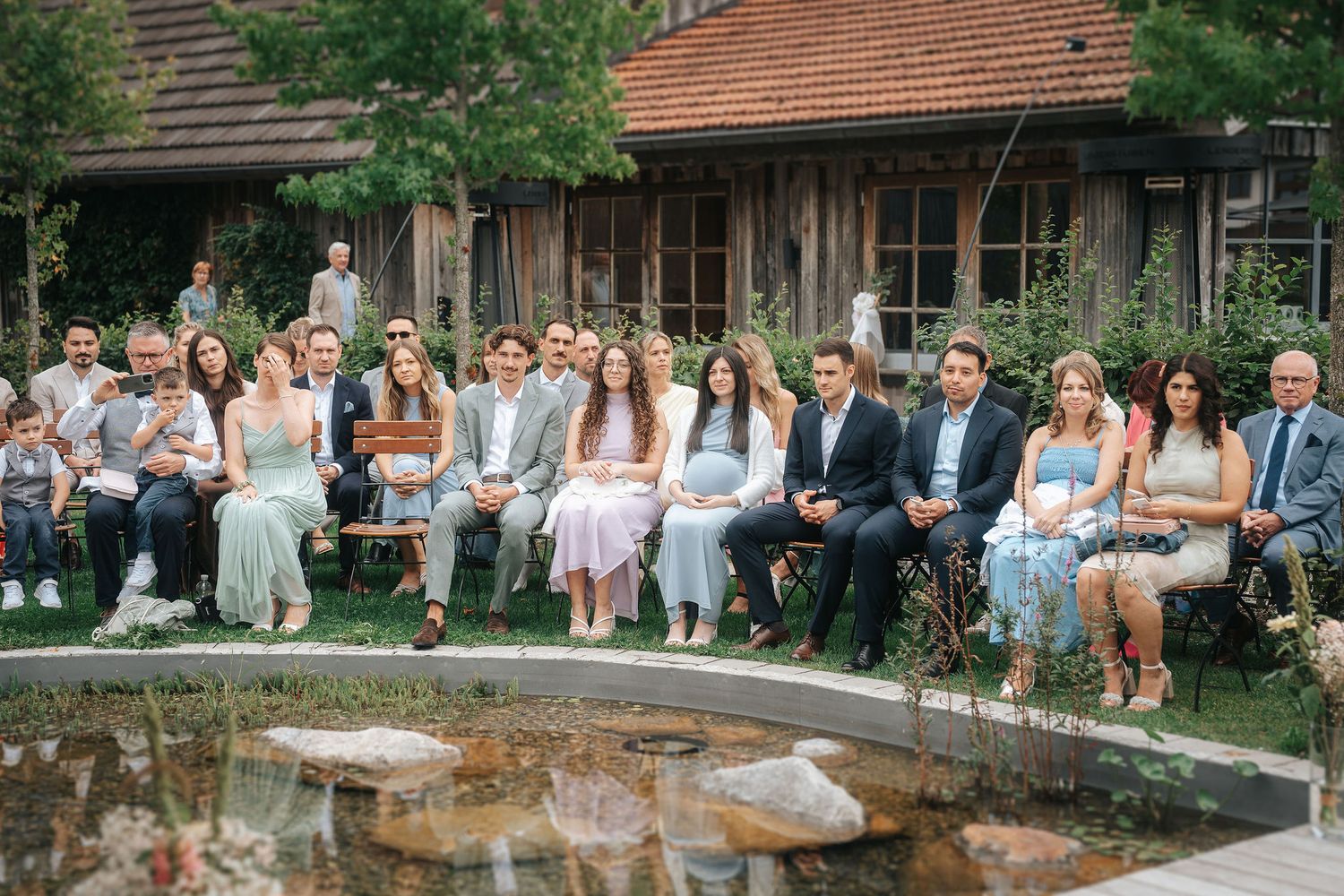 Wedding guests in elegant attire sit on chairs in a garden watching an outdoor ceremony beside a small pond and a rustic wooden building.