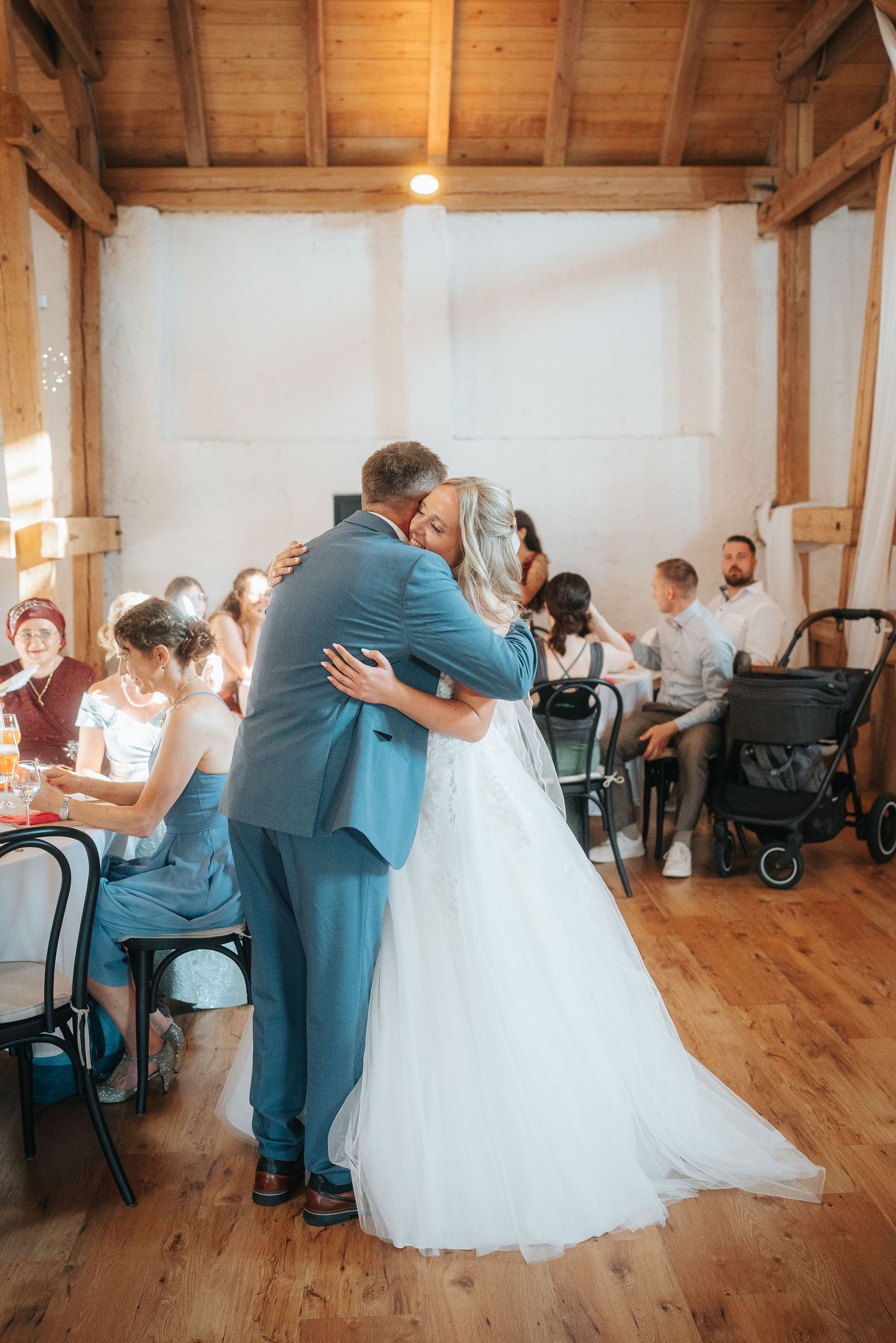 Bride in a white tulle gown embraces a man in a blue suit on a wooden floor while wedding guests watch from tables in a rustic barn.