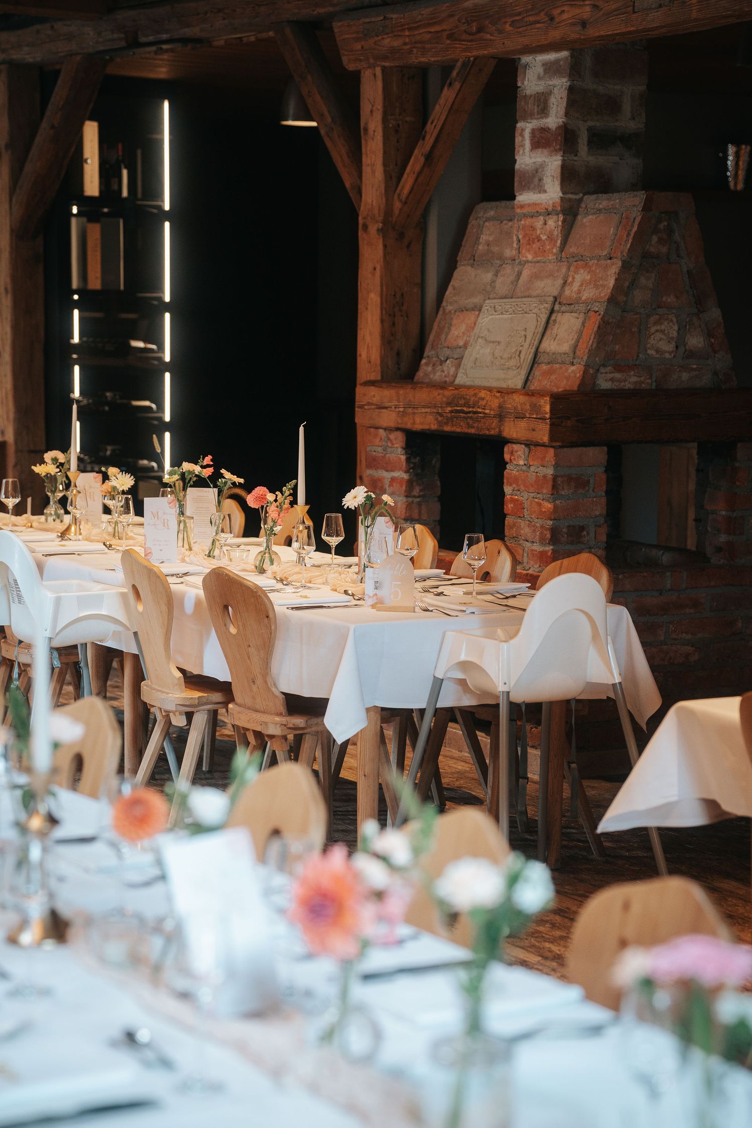 Rustic wedding reception hall with white-draped tables, wooden chairs, fresh flower centerpieces and a brick fireplace under timber beams.