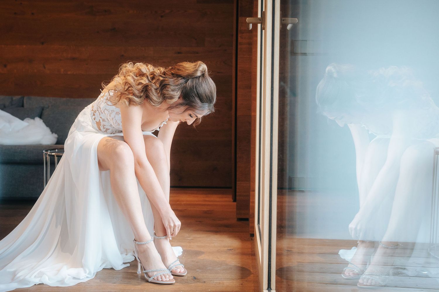 Bride in a flowing white wedding gown sits by a window, bending forward to fasten her strappy heeled sandals before the ceremony.