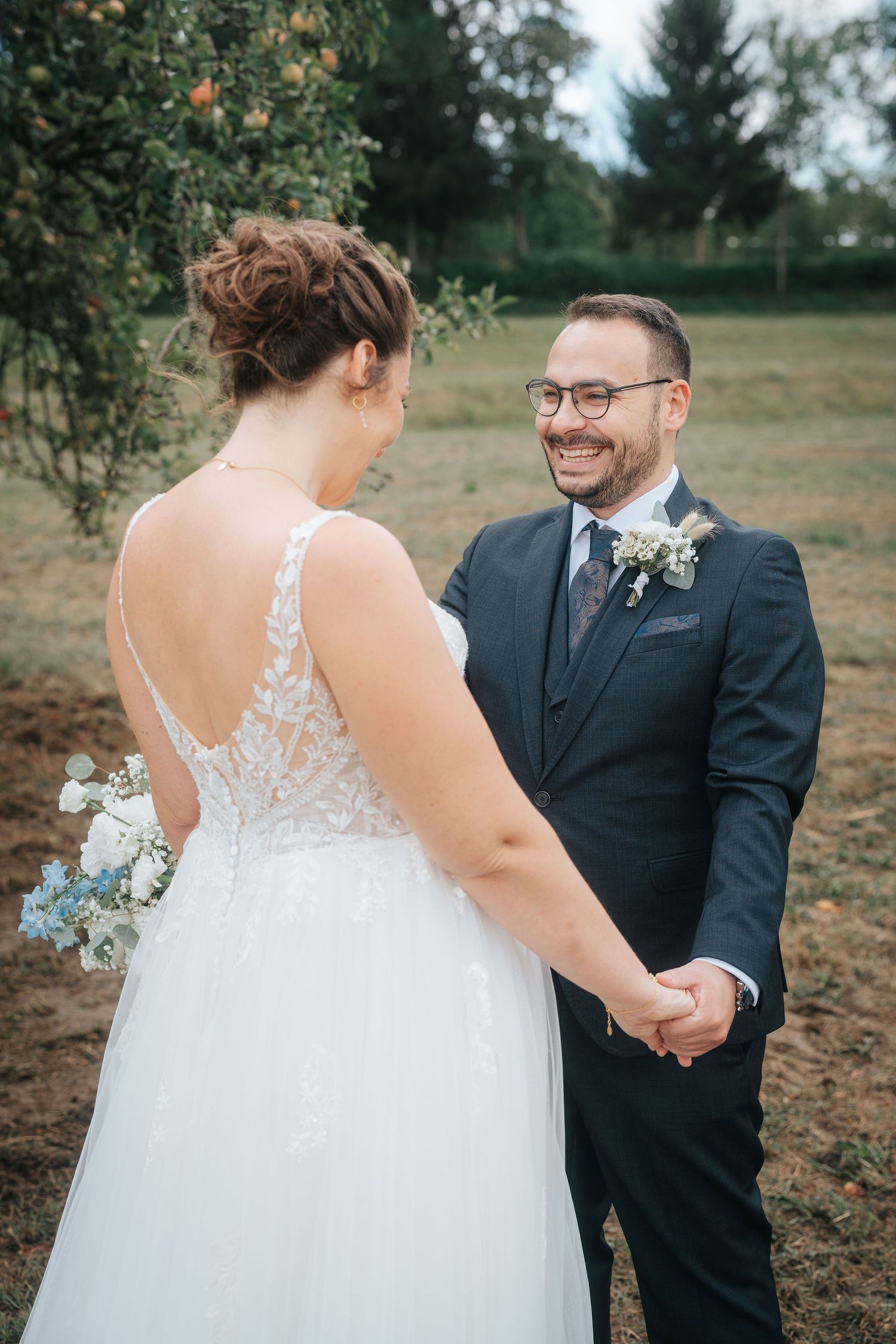 Smiling groom in a navy suit and glasses holds hands with a bride in a lace wedding dress during an outdoor first look.