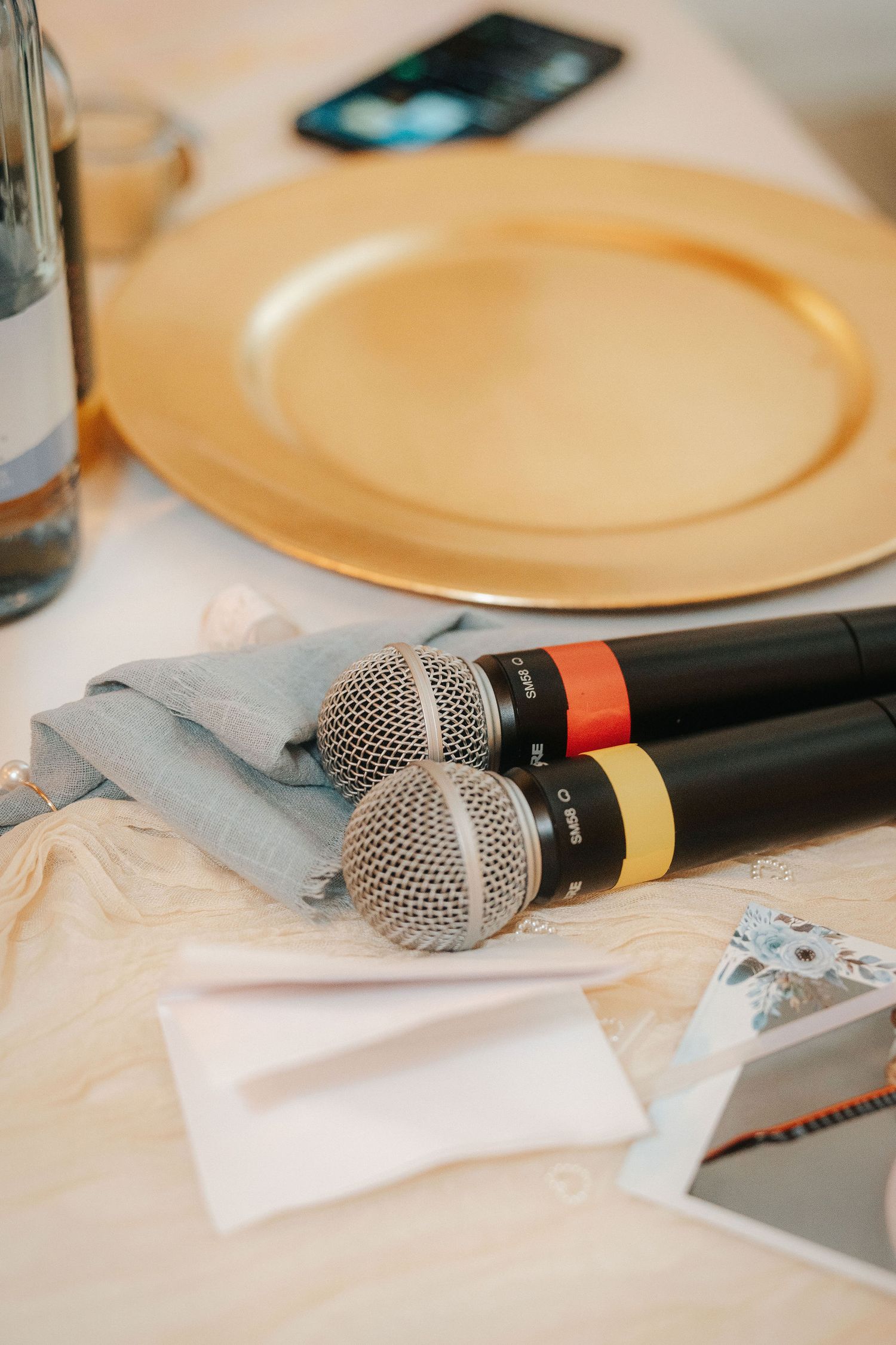 Two black wireless microphones with red and yellow bands rest on a table beside a gold charger plate, gray napkin and place cards.