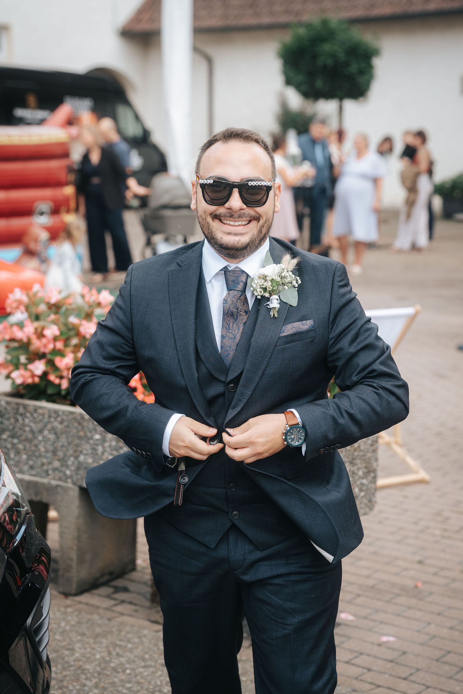 Smiling groom in sunglasses and a navy three-piece suit with a floral boutonniere adjusts his jacket at an outdoor wedding gathering.