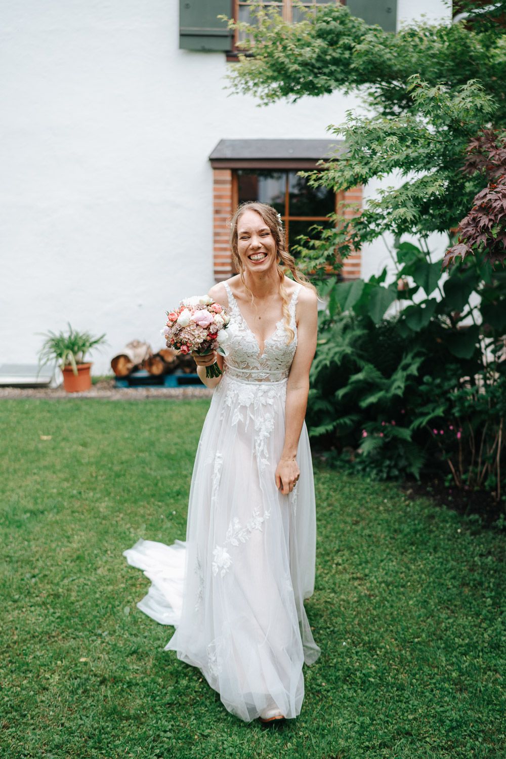 A person wearing a flowing white lace wedding gown stands in a garden with green foliage.