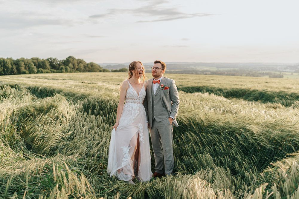 A couple in wedding attire stands together in a scenic golden wheat field during sunset.