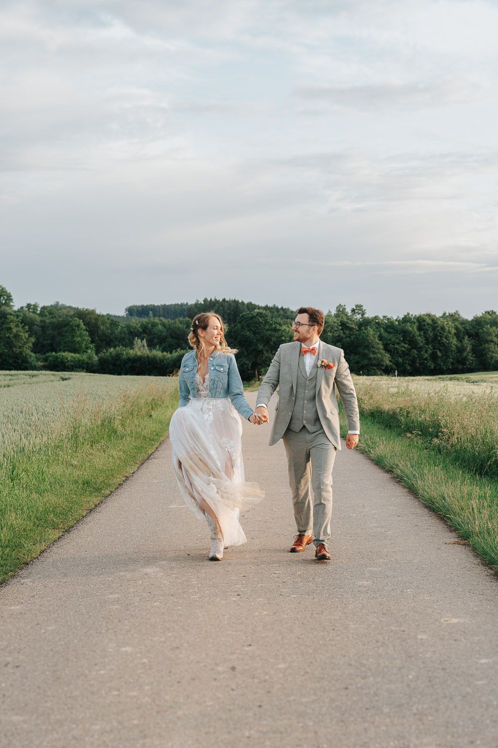 A bride and groom walk hand in hand down a country path surrounded by meadows and trees on a cloudy summer day.