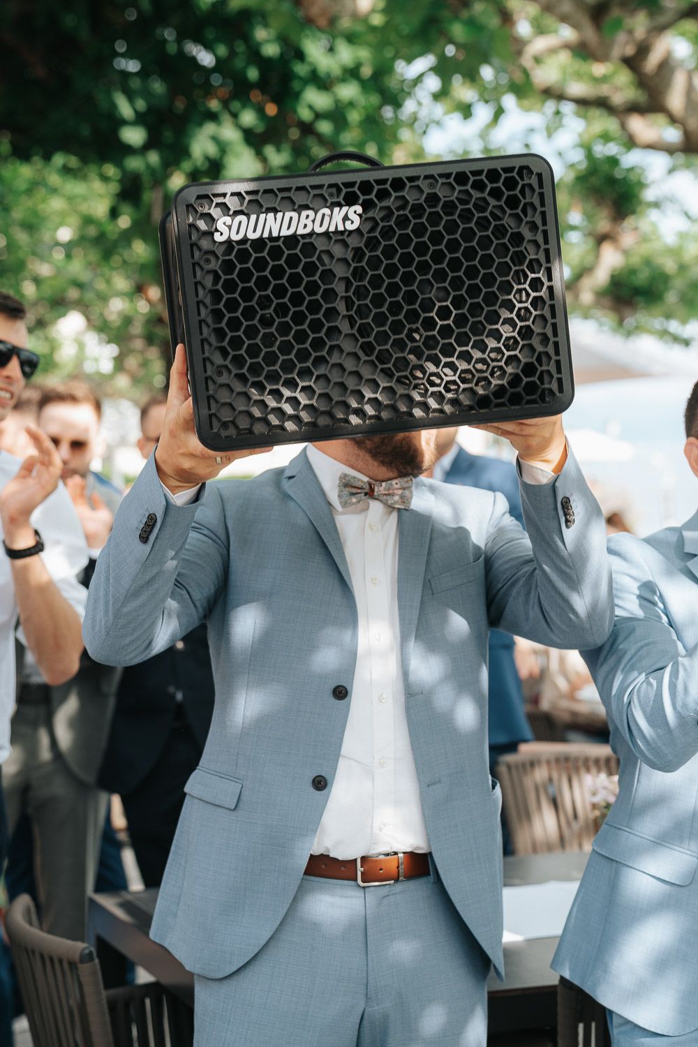 Wedding guest in light blue suit holds up a black Soundboks speaker in front of them during outdoor celebration.