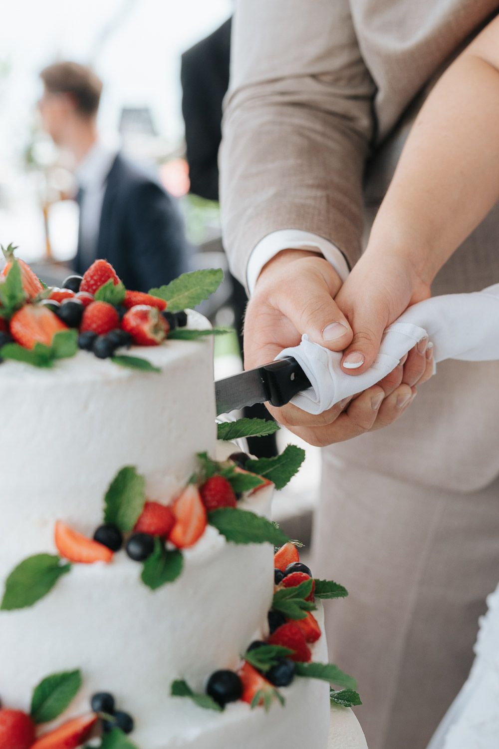 A person cutting into a white wedding cake decorated with fresh strawberries and mint leaves.