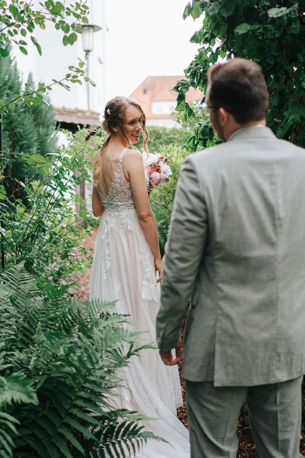 A romantic garden wedding moment with an elegant lace dress and grey suit among lush green ferns and foliage.