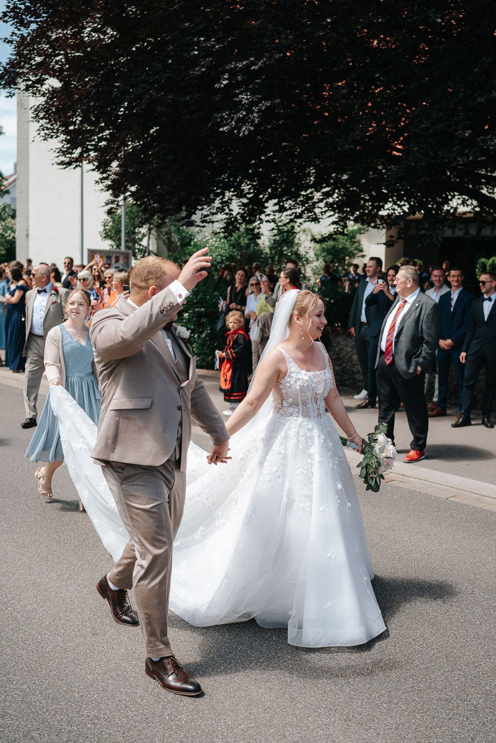 A wedding party walks together on a paved road beneath a large tree during a sunny outdoor celebration.