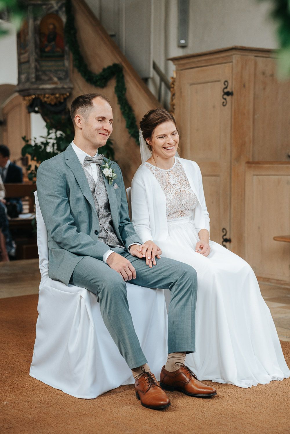 Newlyweds sit together on white steps during wedding photography session in bright sunlit indoor venue.