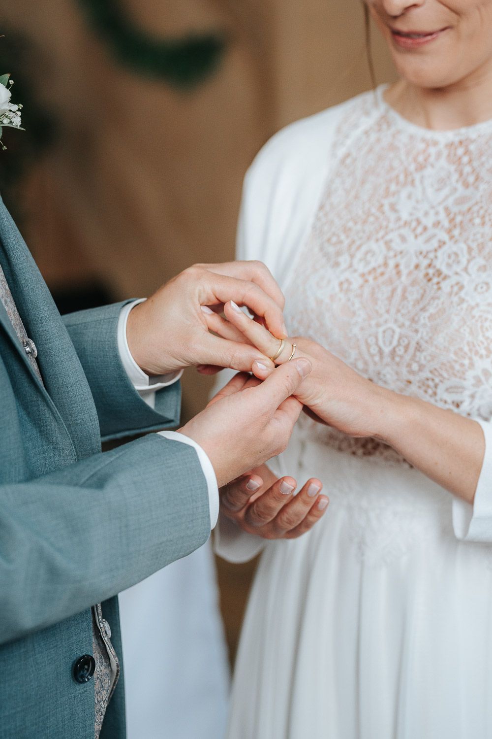 Close-up of exchanging wedding rings during ceremony, with groom in grey suit and bride in white lace dress.