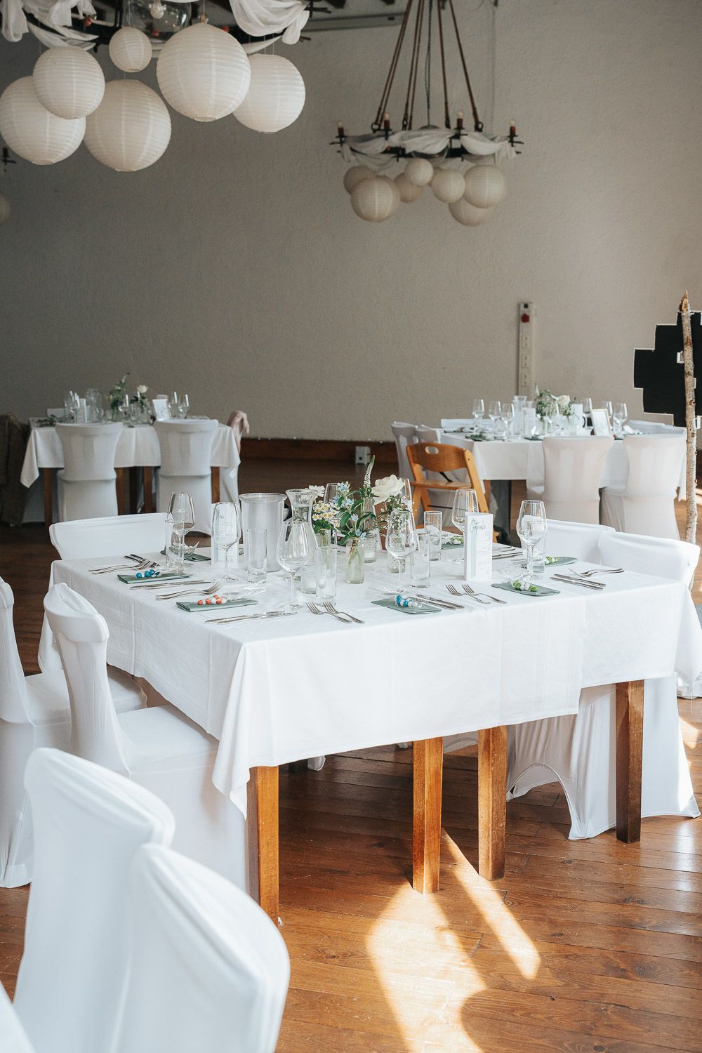 Elegant wedding reception table with white linens, chair covers, and globe pendant lights in a bright dining room.