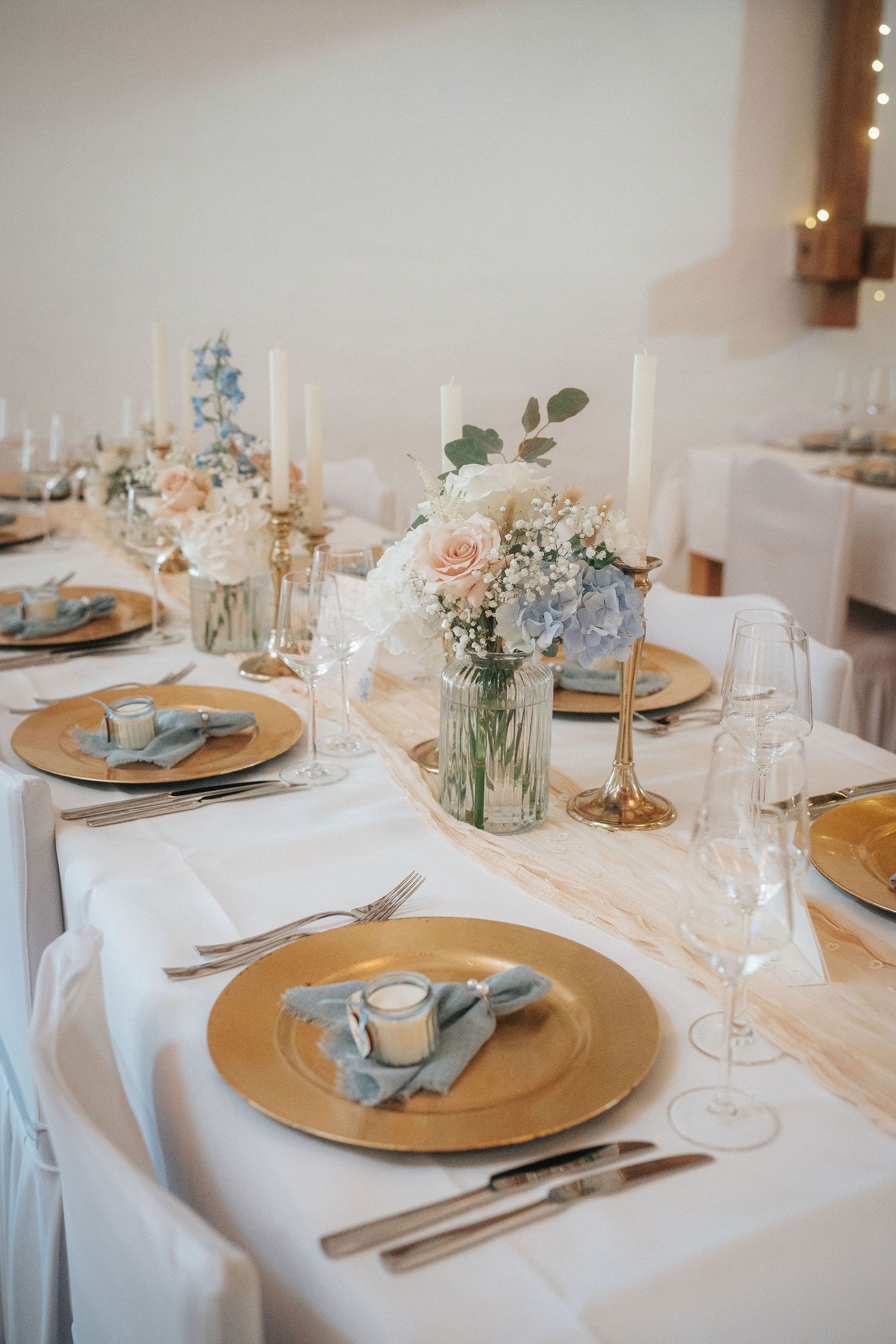 Elegant wedding table setting with gold chargers, dusty blue napkins, pastel floral centerpieces, tall candles, and crystal glassware.