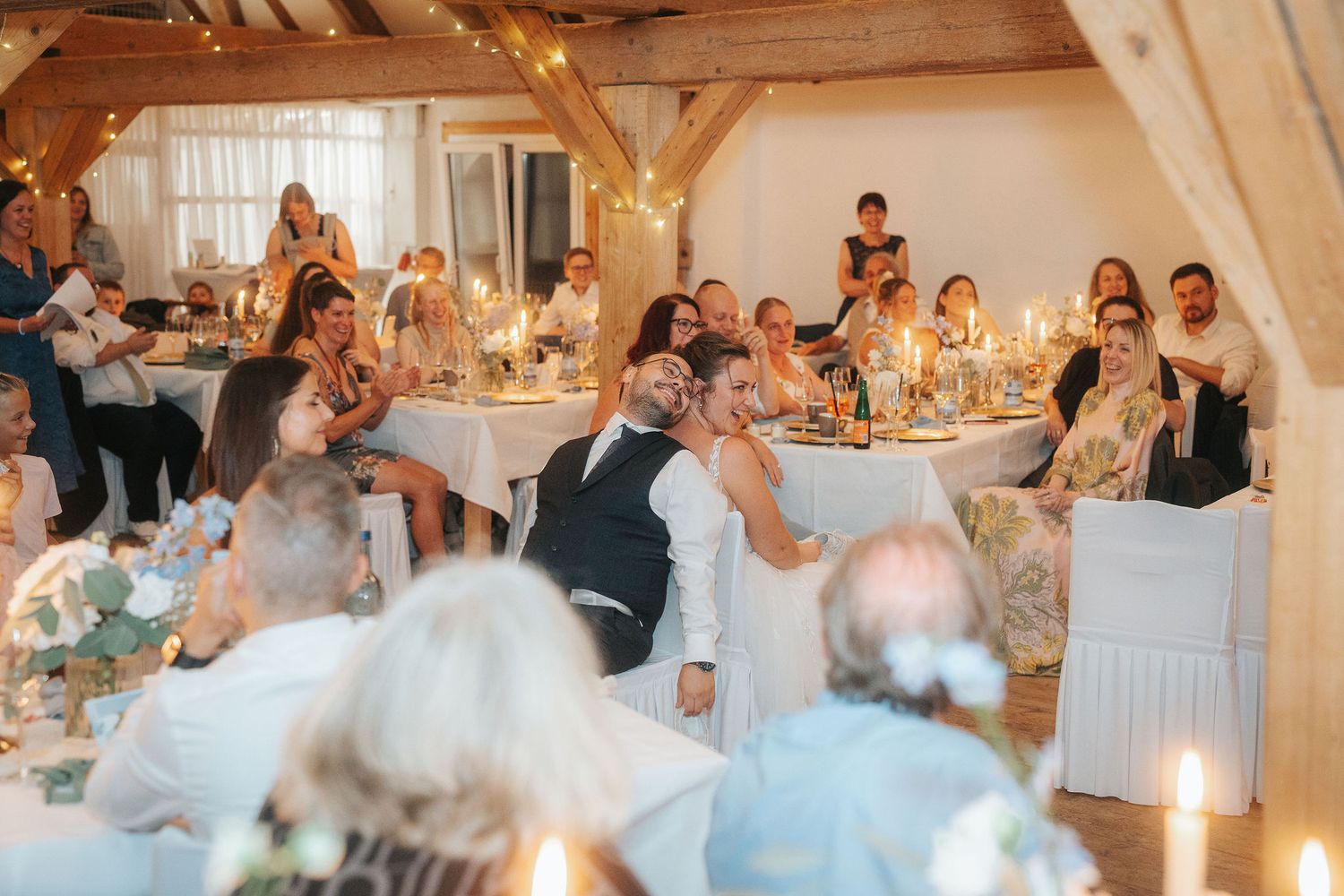 Wedding guests seated at round tables in a rustic wooden barn venue, smiling and laughing together by candlelight during a reception.