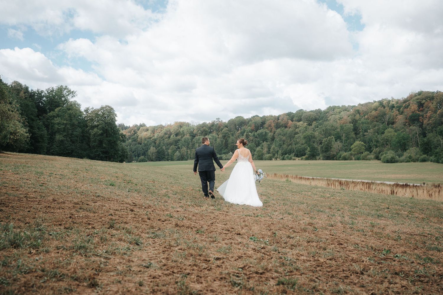 Bride in a white gown and groom in a dark suit hold hands and walk across a grassy field toward a tree-lined hillside under a cloudy sky.
