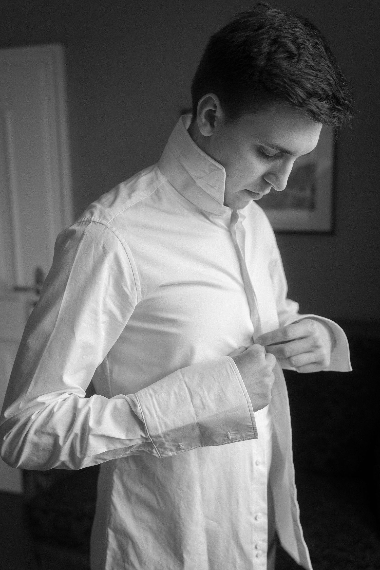 Black and white photo of a man in a white dress shirt with a high collar fastening his cuff while getting dressed by a window.