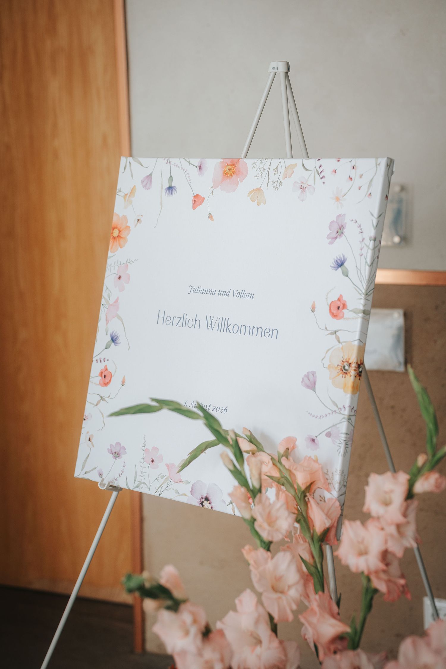 Floral wedding welcome sign reading Herzlich Willkommen on an easel, framed by watercolor wildflowers and peach gladiolus blooms.