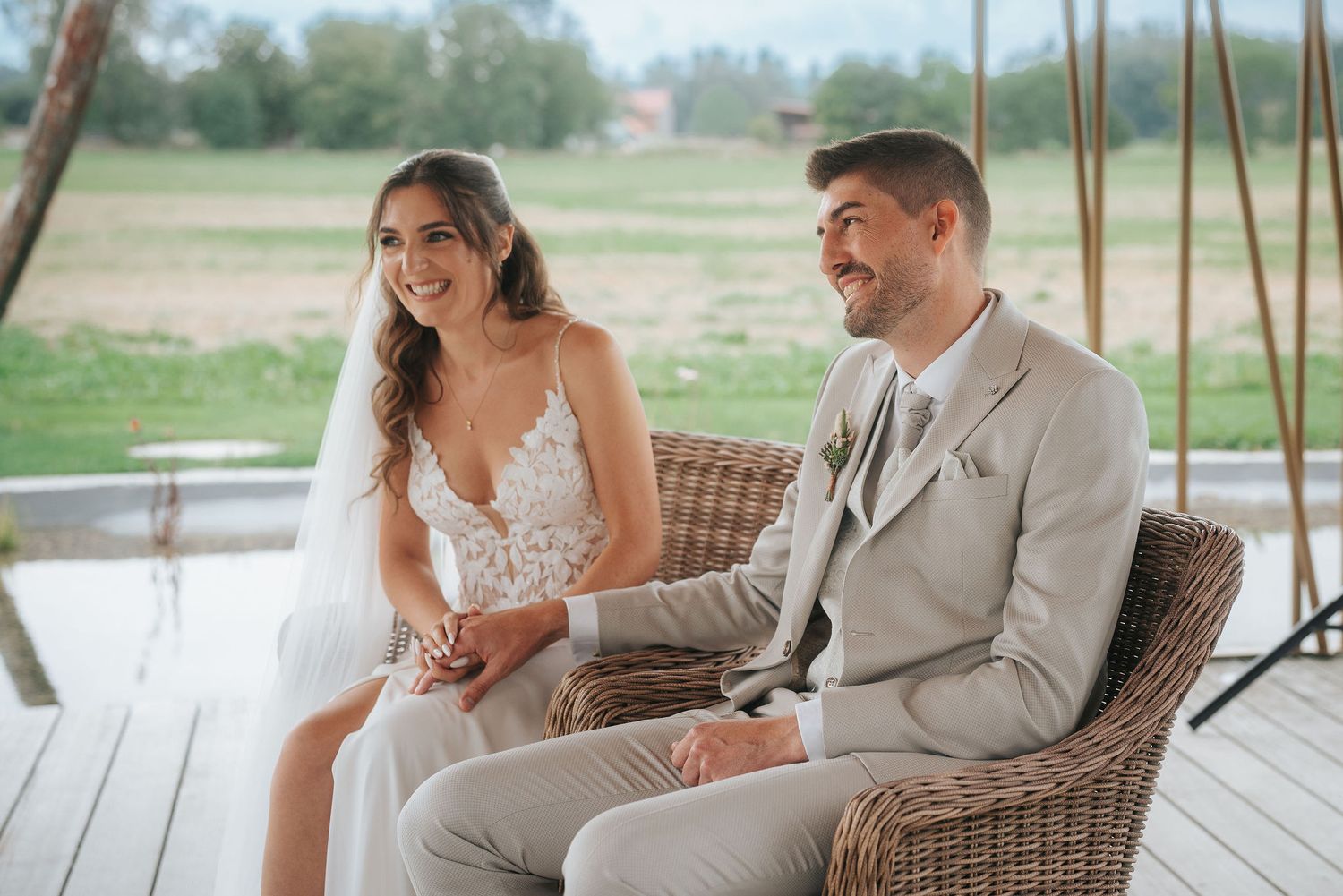 Smiling bride in a lace wedding dress and veil sits beside a groom in a beige suit on wicker chairs during an outdoor ceremony.