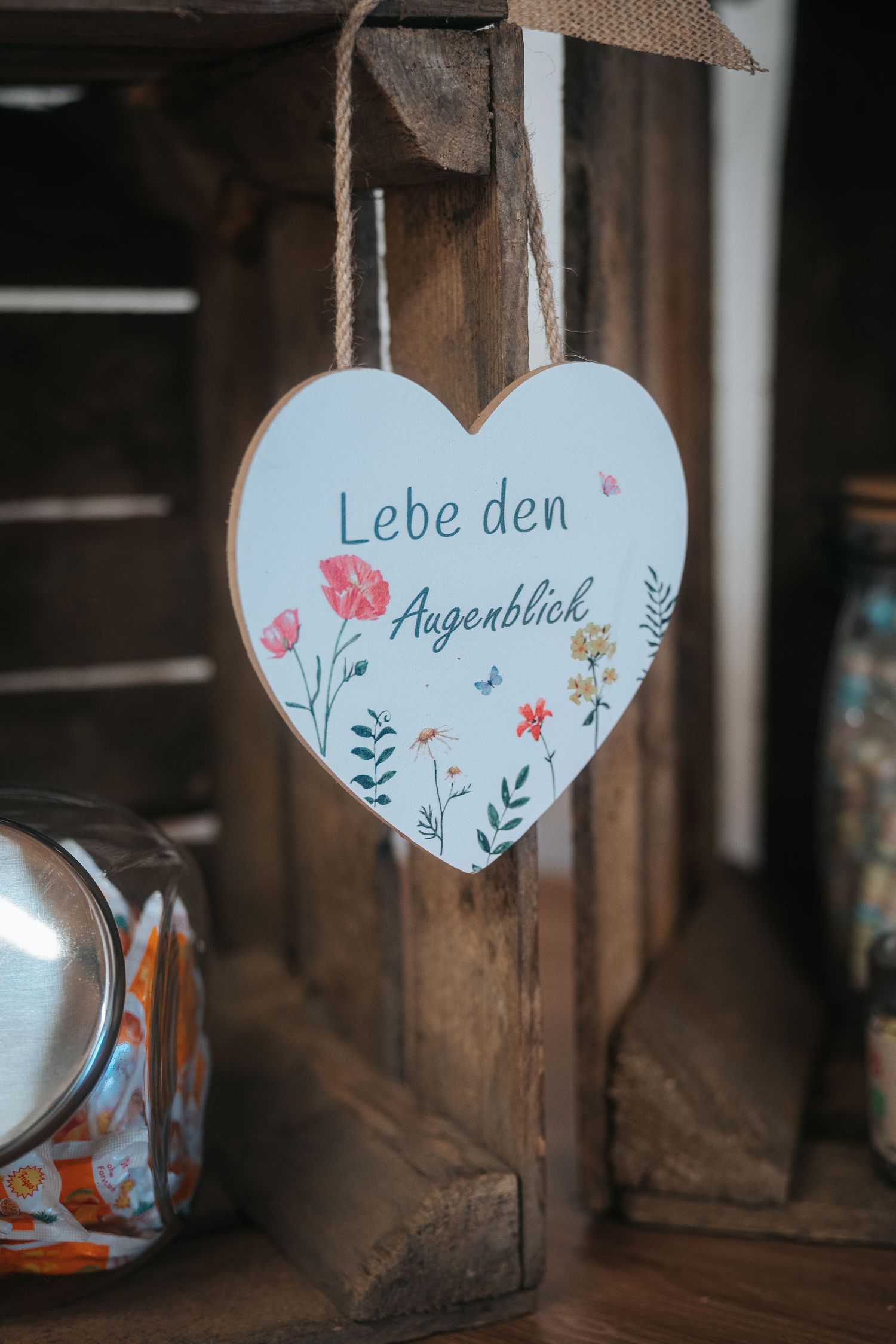Hanging heart-shaped wooden sign reading Lebe den Augenblick with colorful wildflower illustrations, displayed on a rustic wooden crate.