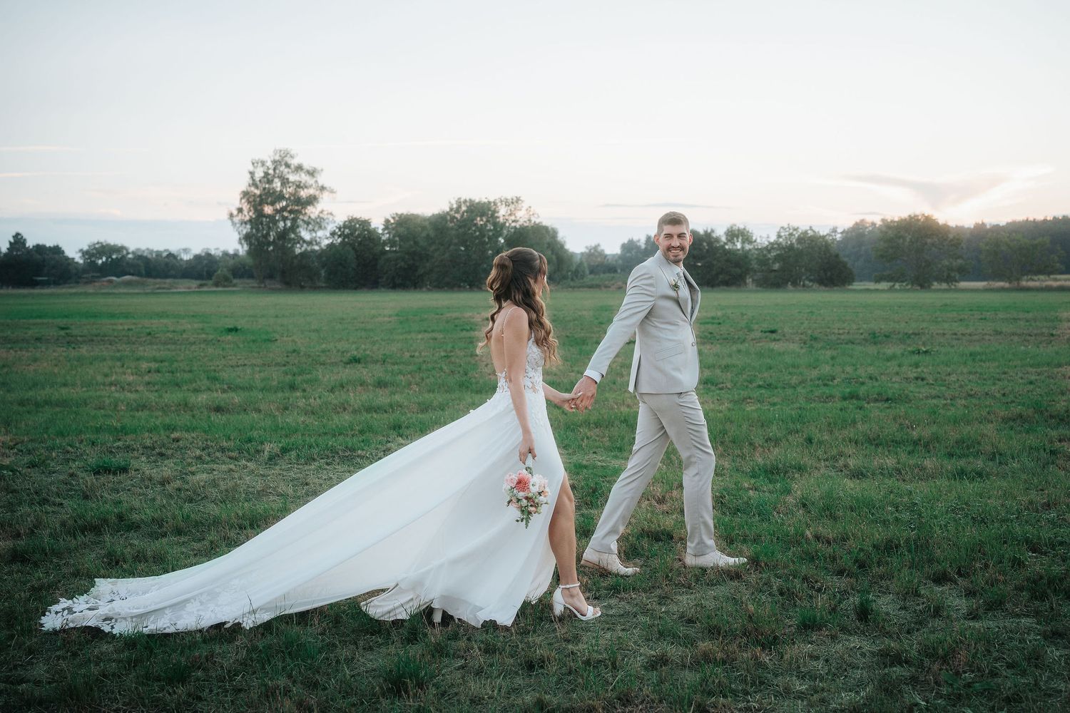 Bride in a flowing white gown and groom in a light gray suit hold hands while walking across a green meadow at sunset.