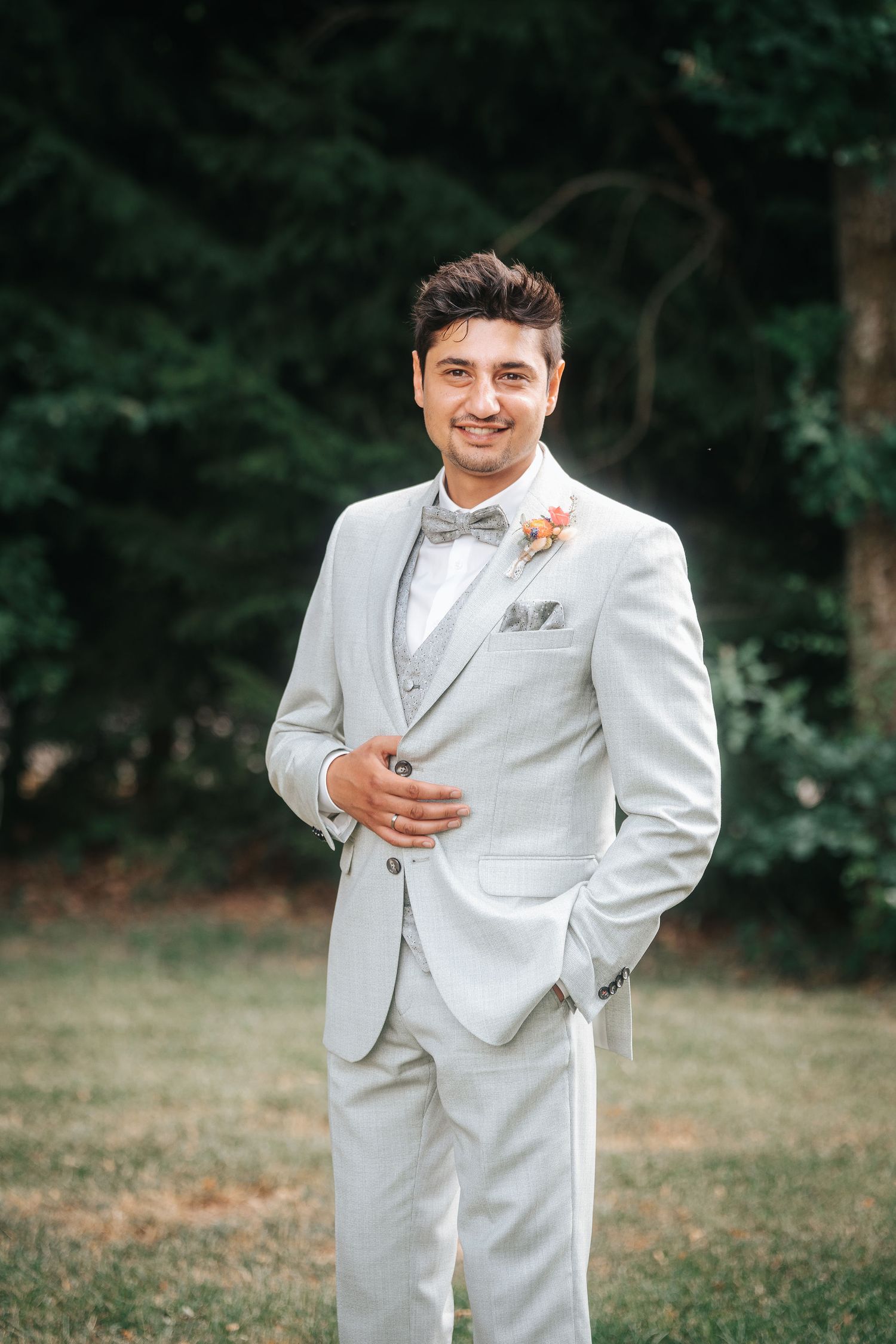 Smiling groom in a light gray three-piece suit with a bow tie and floral boutonniere standing on a grassy lawn in front of green trees.