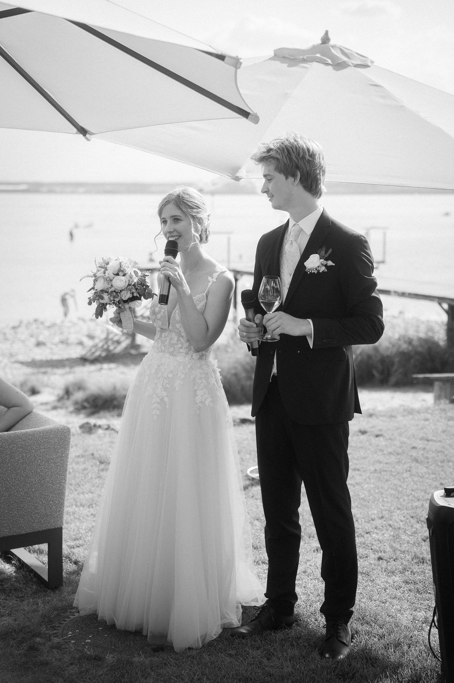 Black and white photo of a bride holding a bouquet and microphone beside a groom with a glass at a lakeside outdoor wedding.