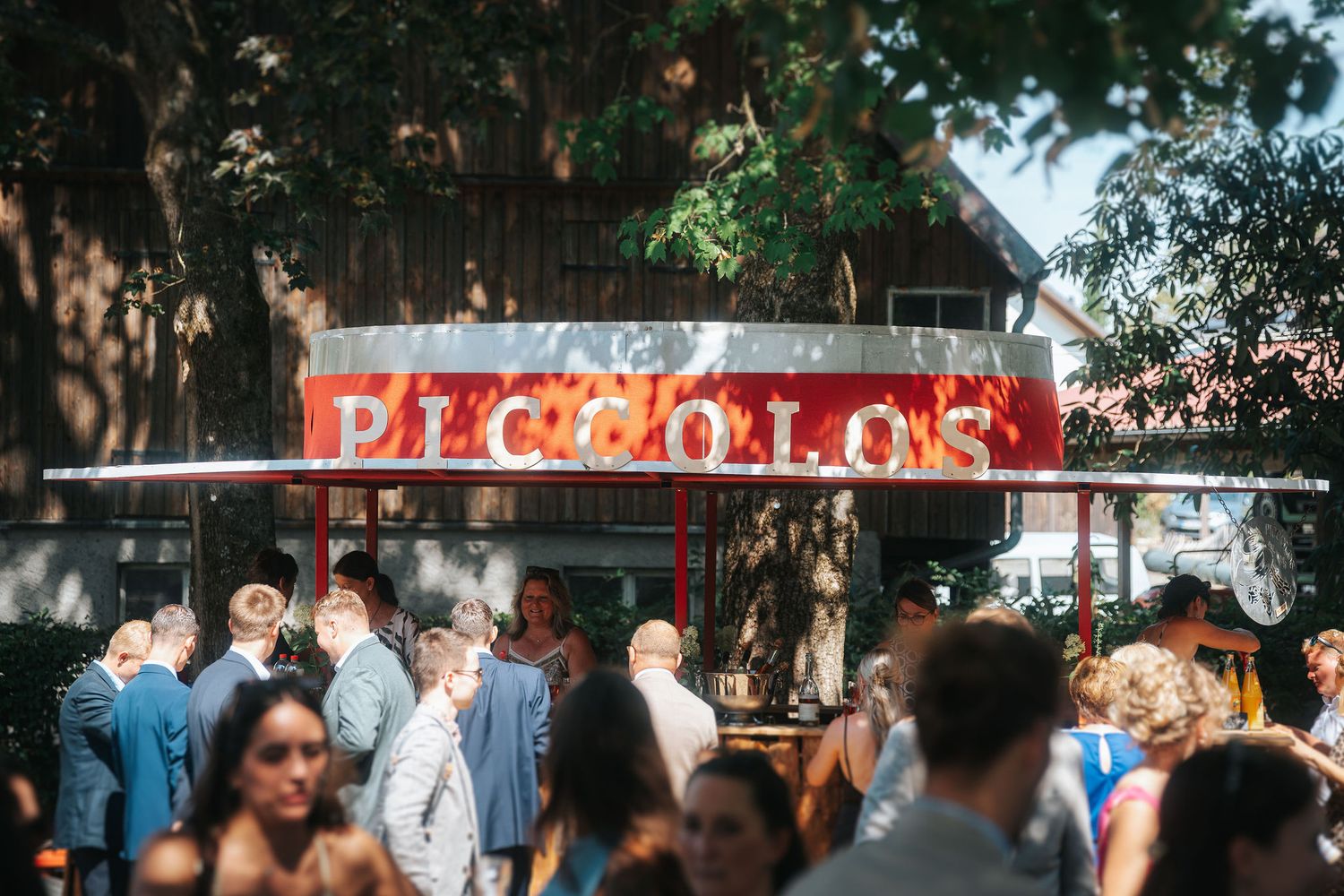 Wedding guests in formal attire gather outdoors beneath trees near a red Piccolo's food stand with illuminated marquee lettering.