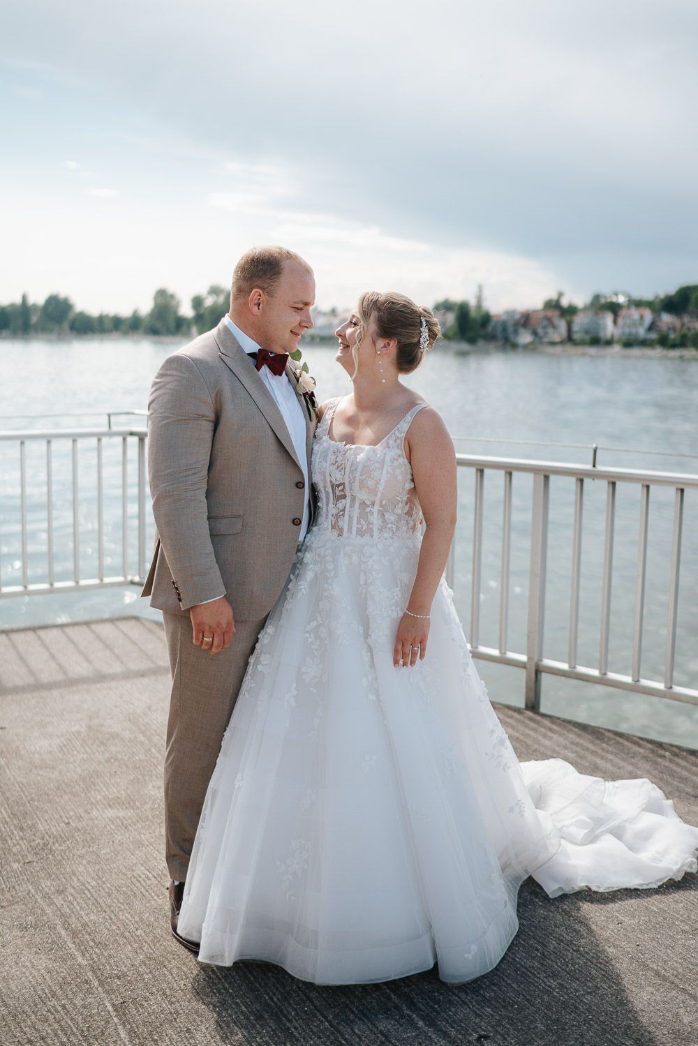 A couple wearing wedding attire embrace on a lakefront pier with a scenic waterfront backdrop on their special day.