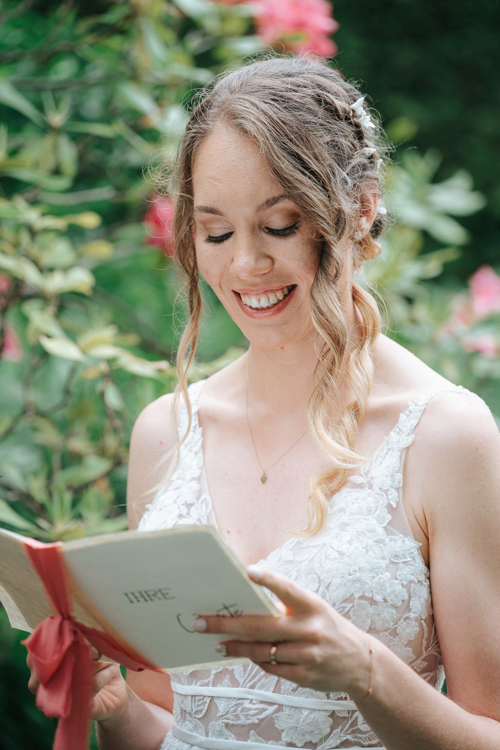 A bride in a white lace dress smiles while reading from a small book in a garden with pink flowers.