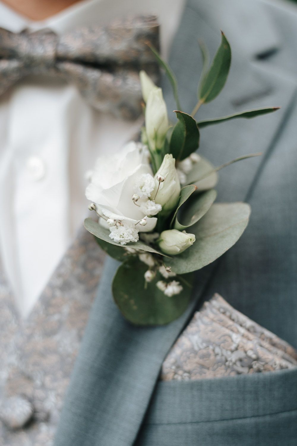 A white floral boutonniere with green leaves pinned to a grey suit pocket.