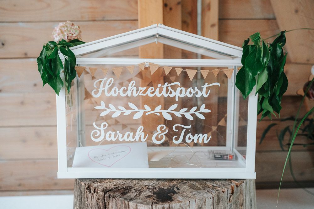 A rustic wedding card box with white lettering spells out 'Hochzeitspost' and names on a clear glass display case.