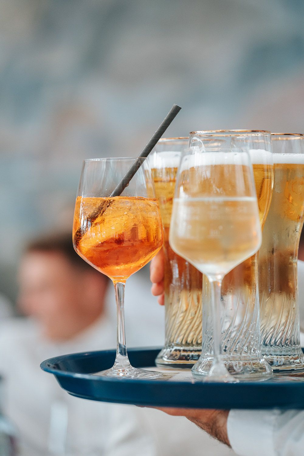 Elegant cocktail glasses with orange aperol spritz and wine being served on a dark blue tray against blurred background.