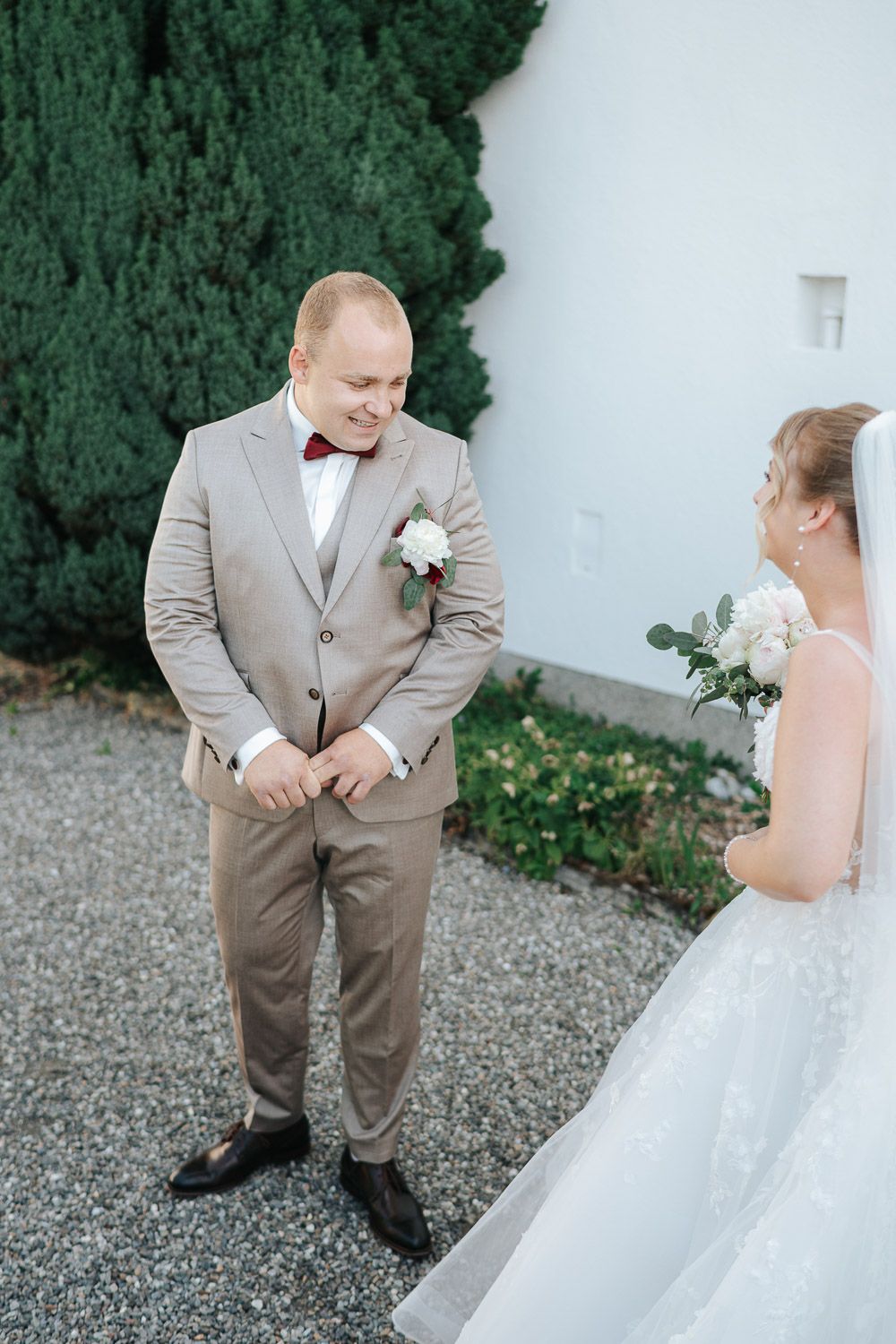 A wedding couple shares a sweet moment outdoors against a backdrop of evergreen trees and white walls.