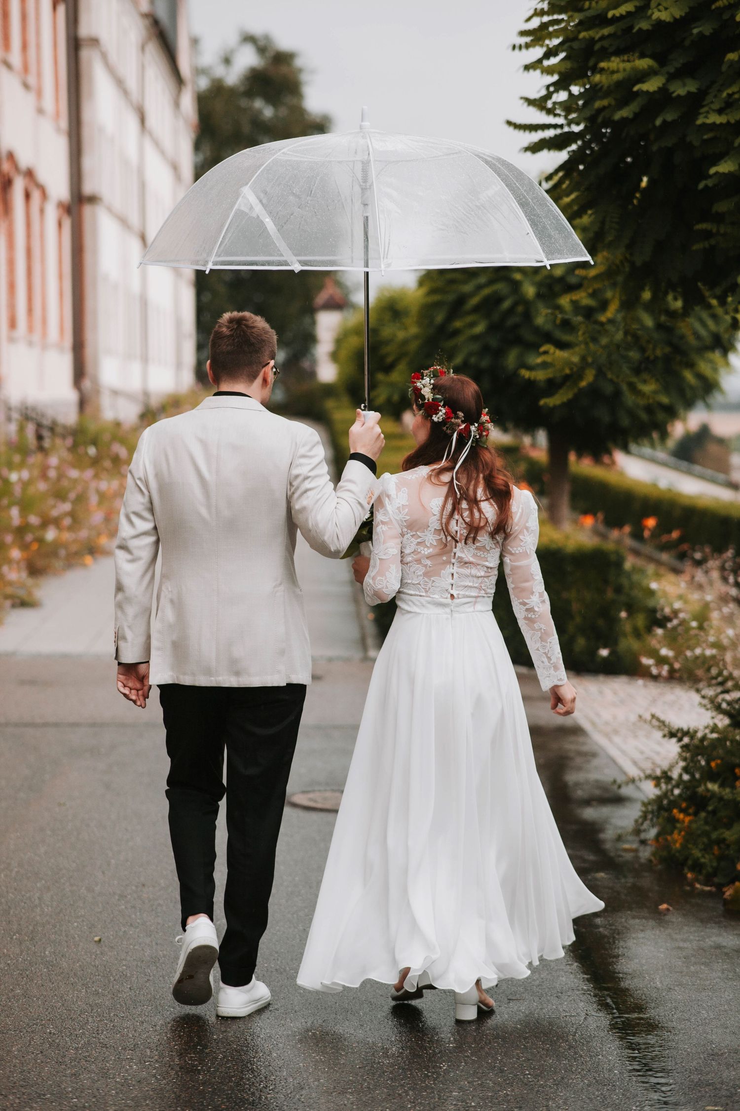 A couple in wedding attire walks together under a clear umbrella on a rainy day.