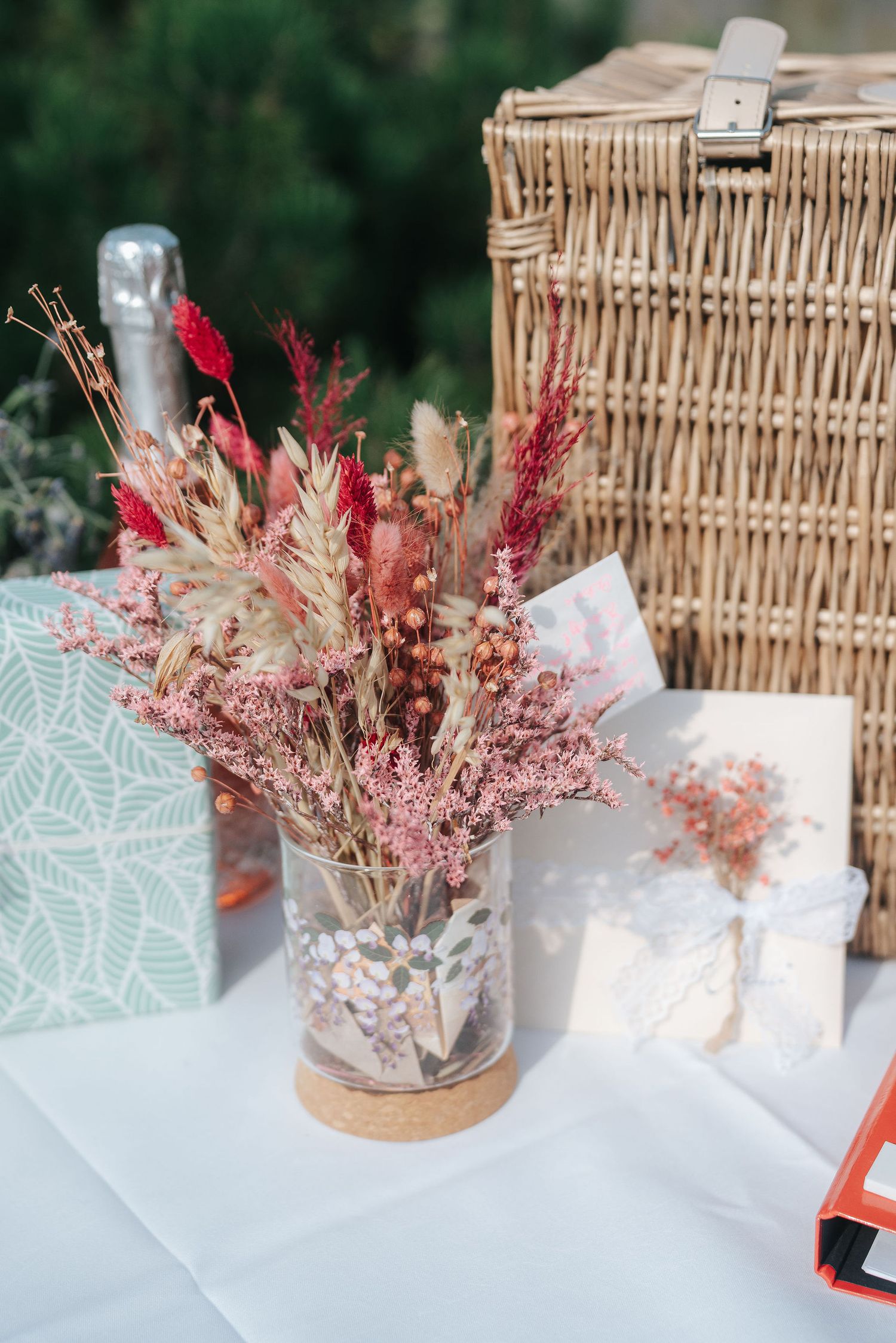 Dried pink and red flower bouquet in a glass vase on a white table beside a wicker picnic basket, champagne bottle and gift card.