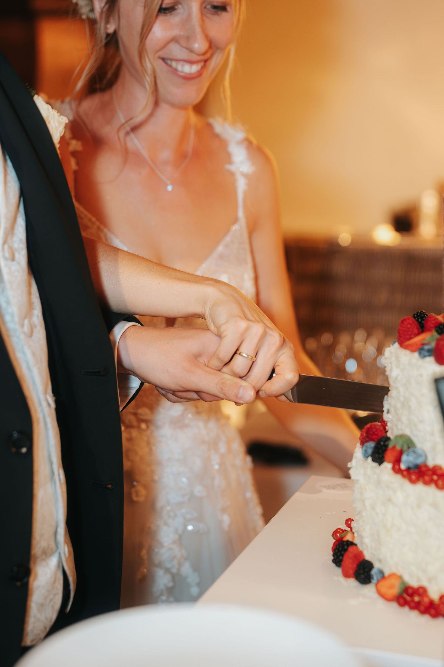 Smiling bride in a lace wedding dress and groom in a vest cut a white berry-topped wedding cake together.