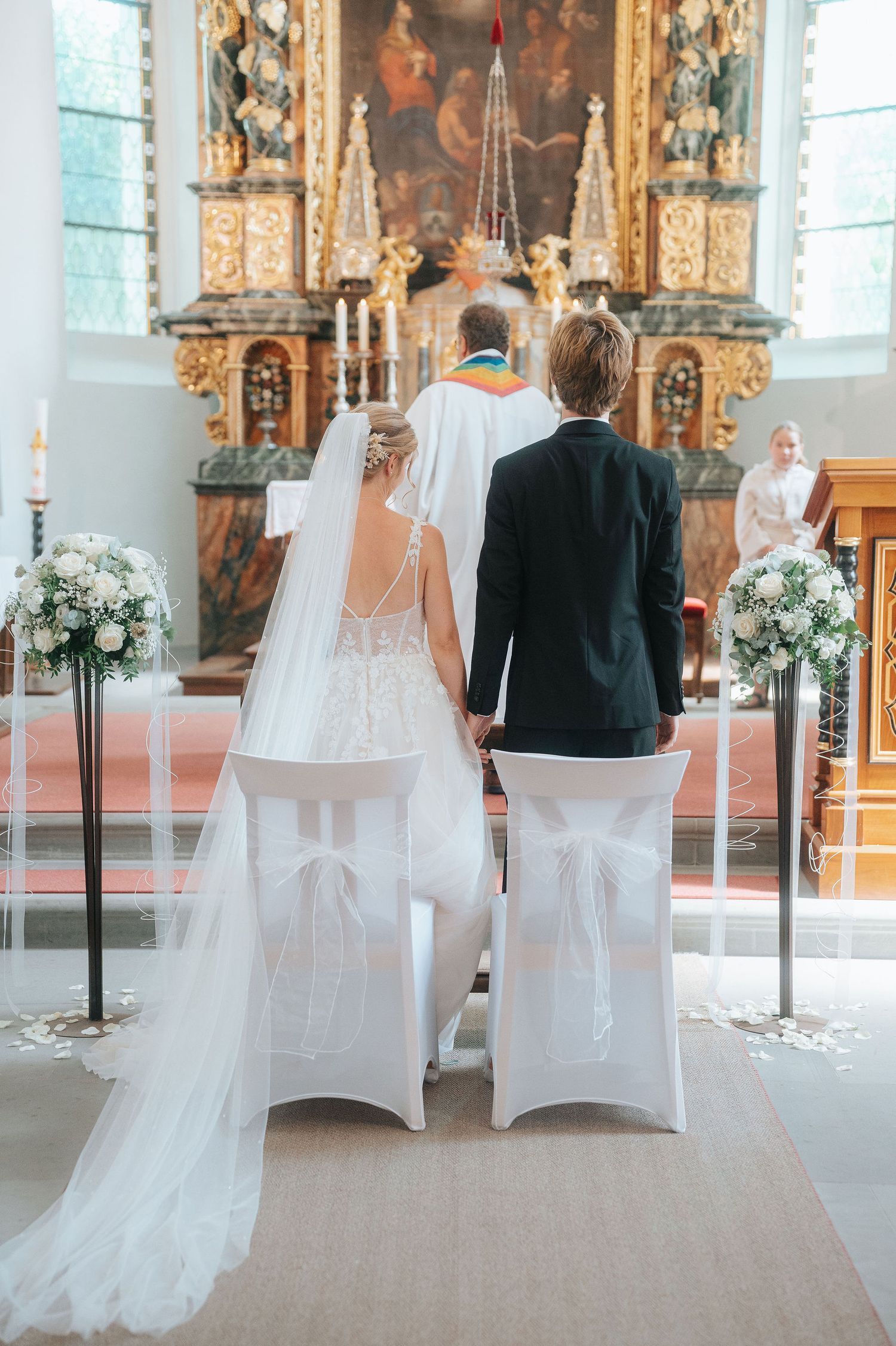 Bride and groom standing at church altar during wedding ceremony with ornate golden decor and white floral arrangements.