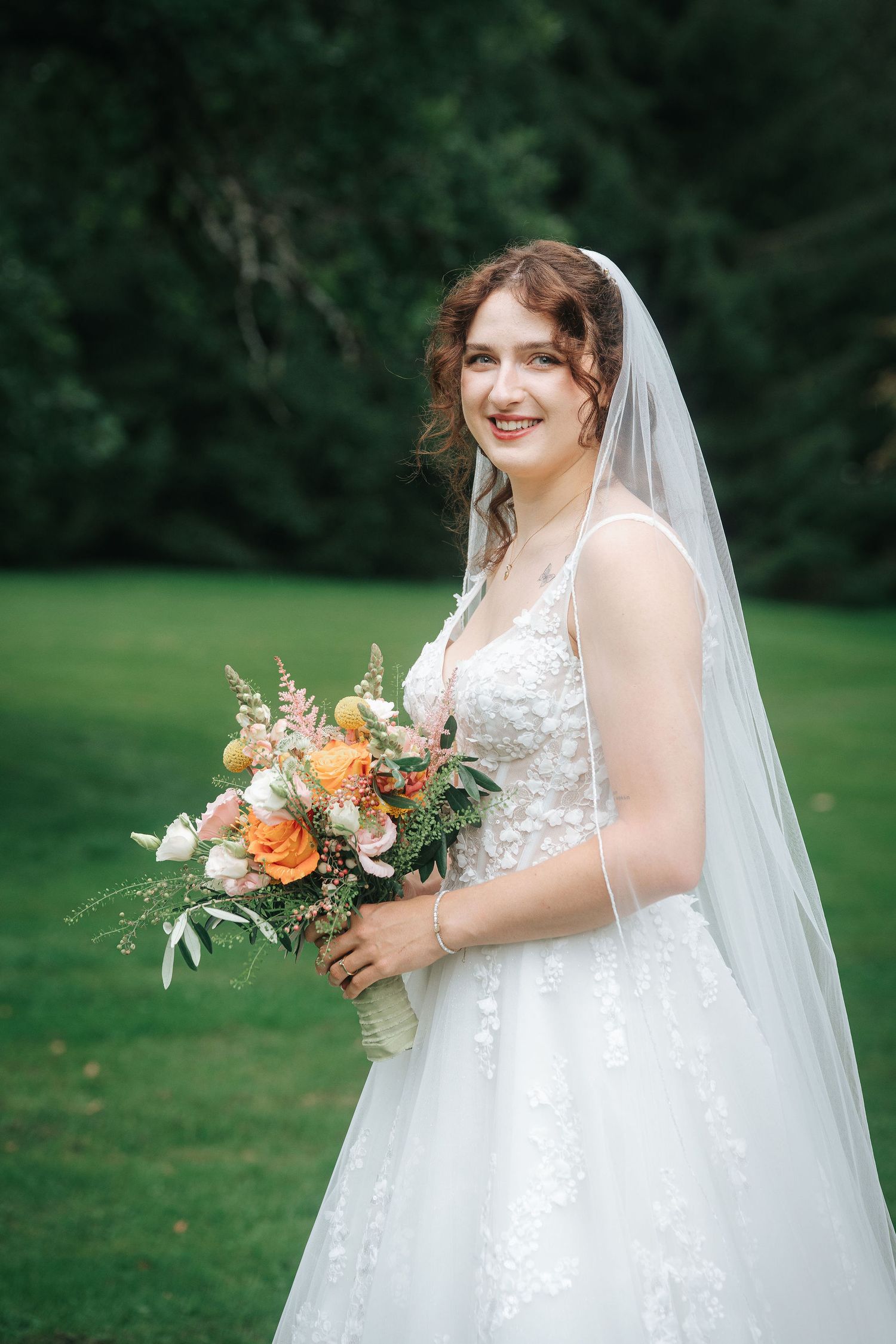 Smiling bride in a lace wedding gown and long veil holds an orange and pink floral bouquet while standing on a green lawn.