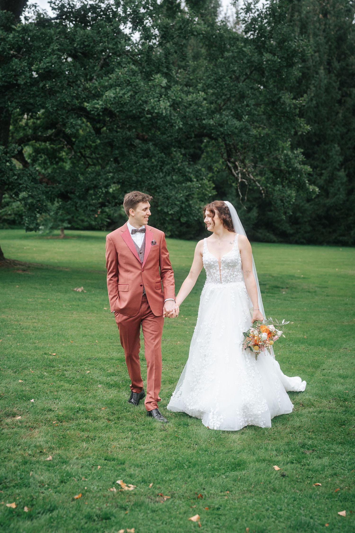 Bride in a white lace wedding gown and groom in a terracotta suit hold hands while walking across a green lawn surrounded by tall trees.