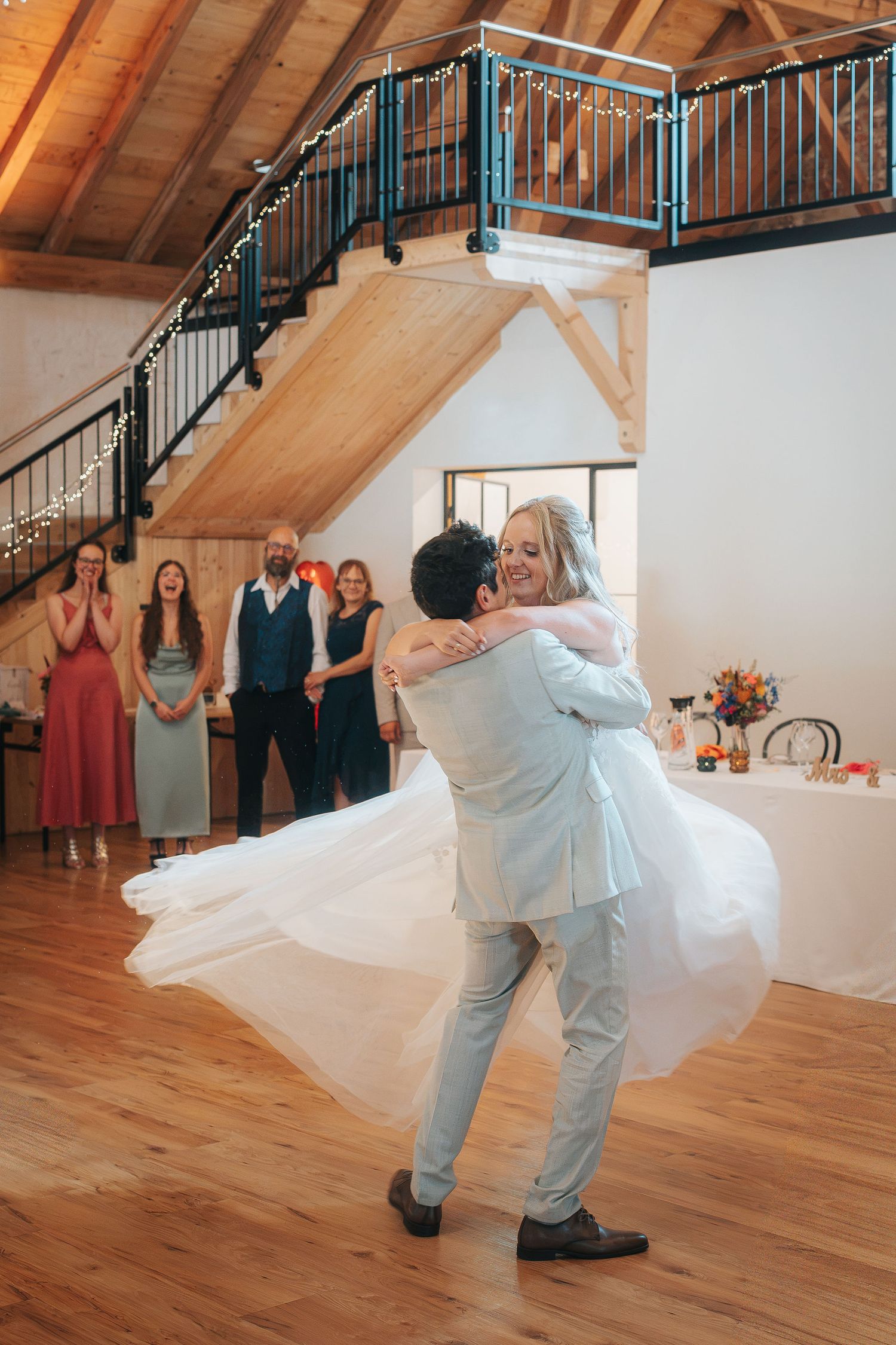 Groom in a light gray suit lifts and spins his bride in a flowing white gown during their first dance as guests watch in a rustic wooden ven
