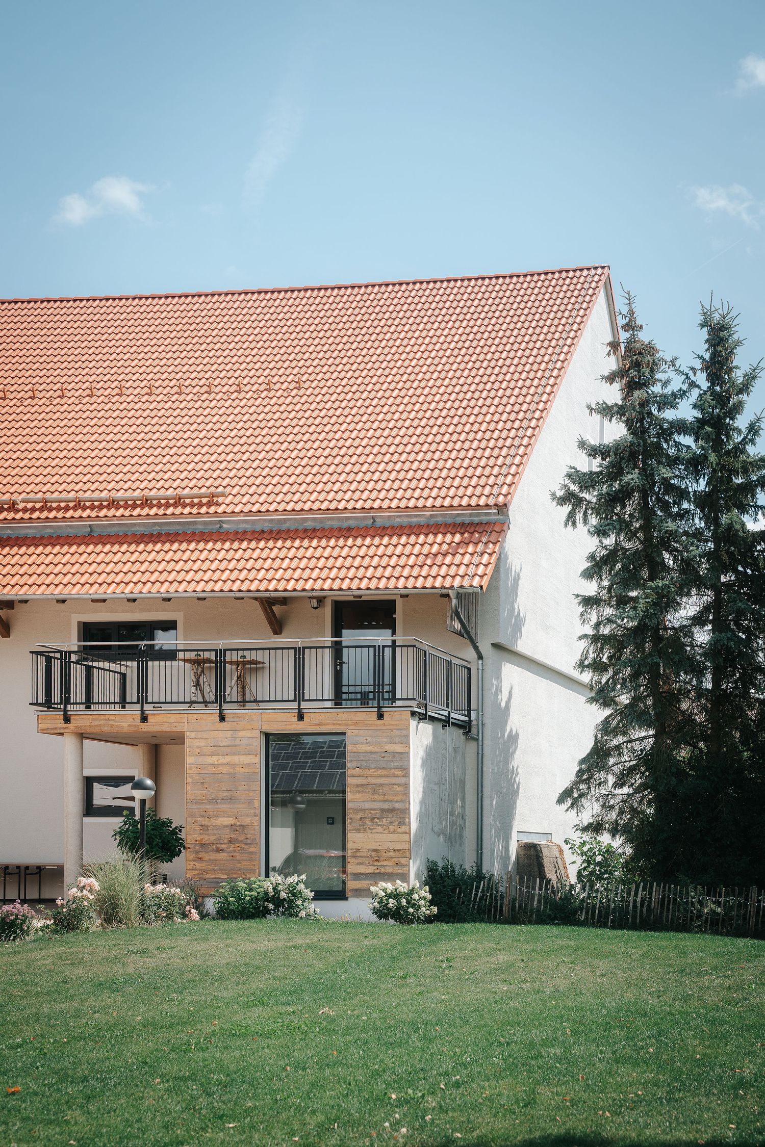 Rustic country house with orange tile roof, wooden balcony and white walls, set beside tall evergreen trees and a green lawn.