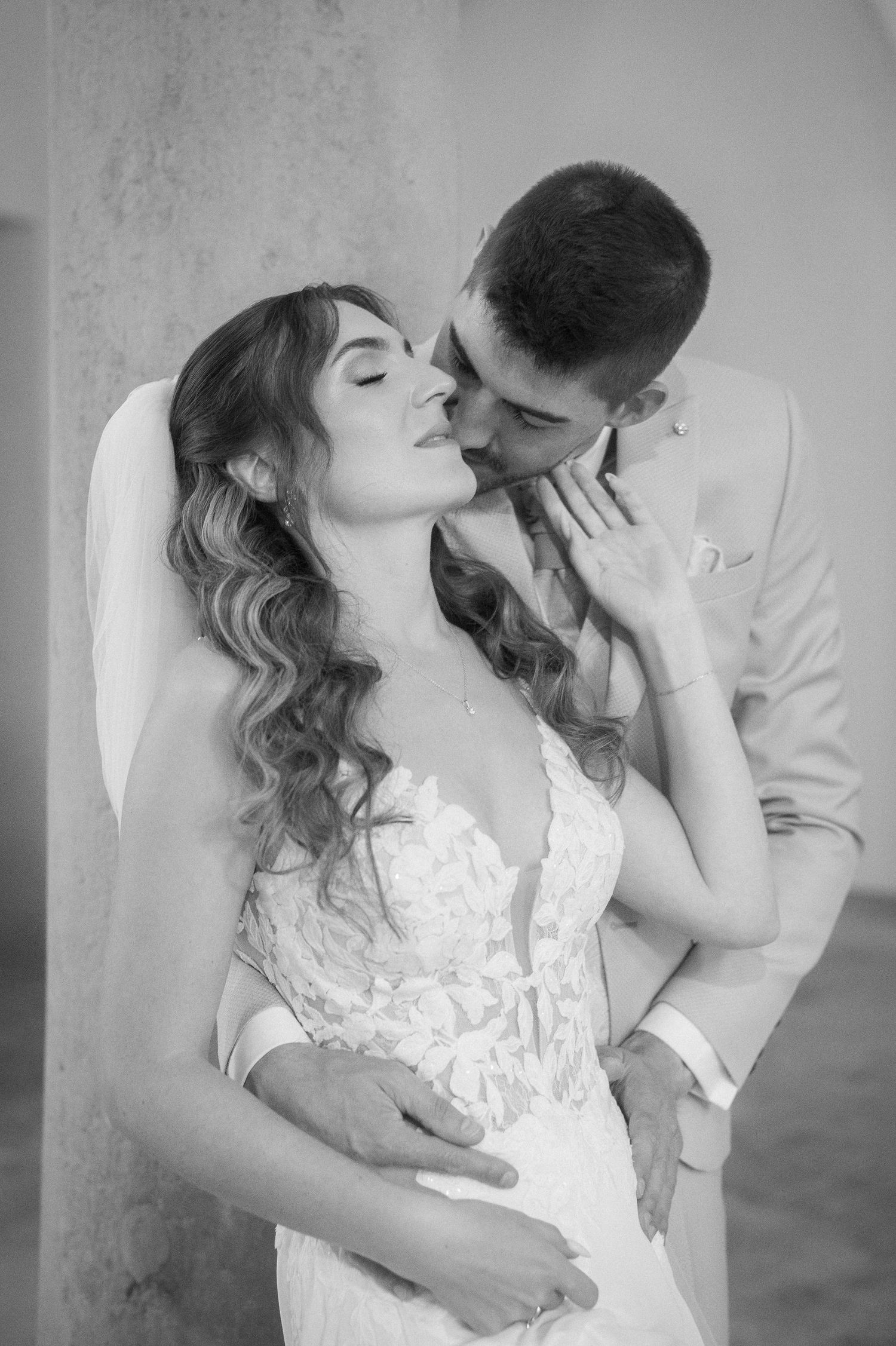 Black and white wedding portrait of a bride in a lace gown with wavy hair and veil being kissed on the cheek by her groom.