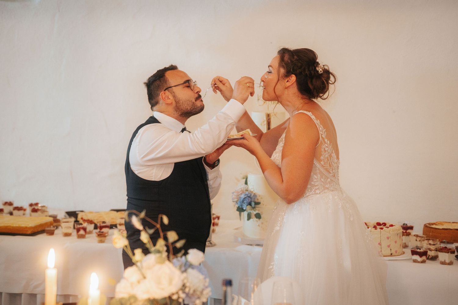 Bride in a white lace gown and groom in a vest feed each other wedding cake at a dessert table with candles and flowers.