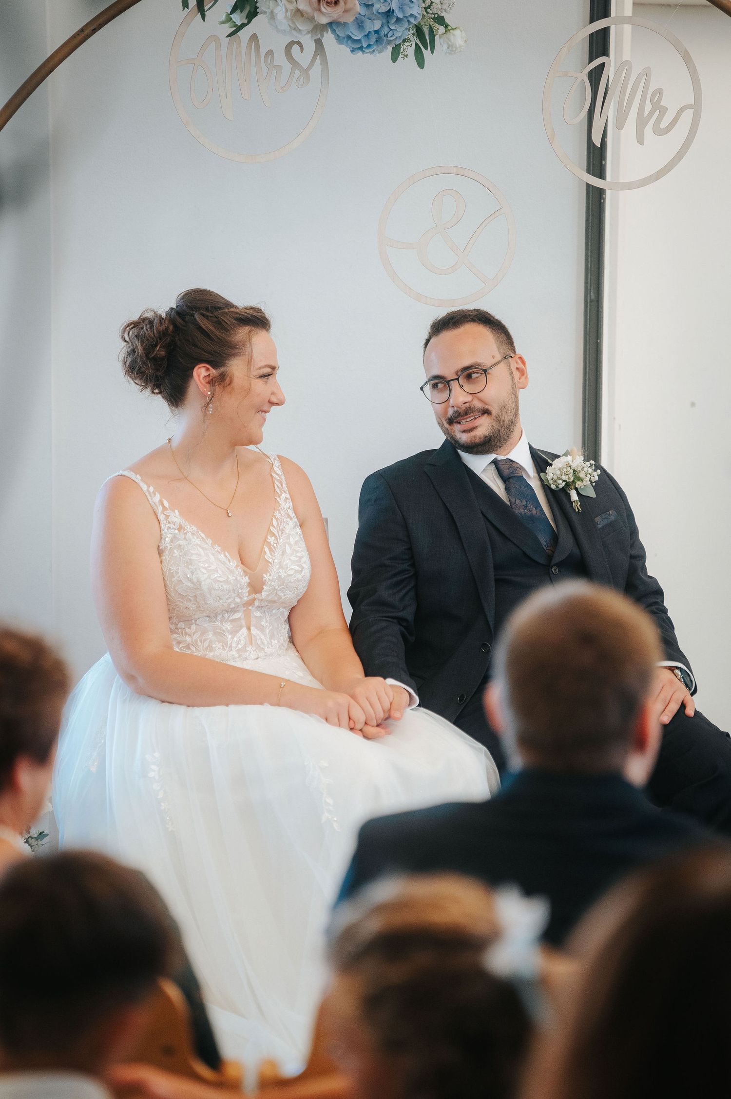 Smiling bride in a lace wedding gown sits beside her glasses-wearing groom in a navy suit during their ceremony as guests look on.