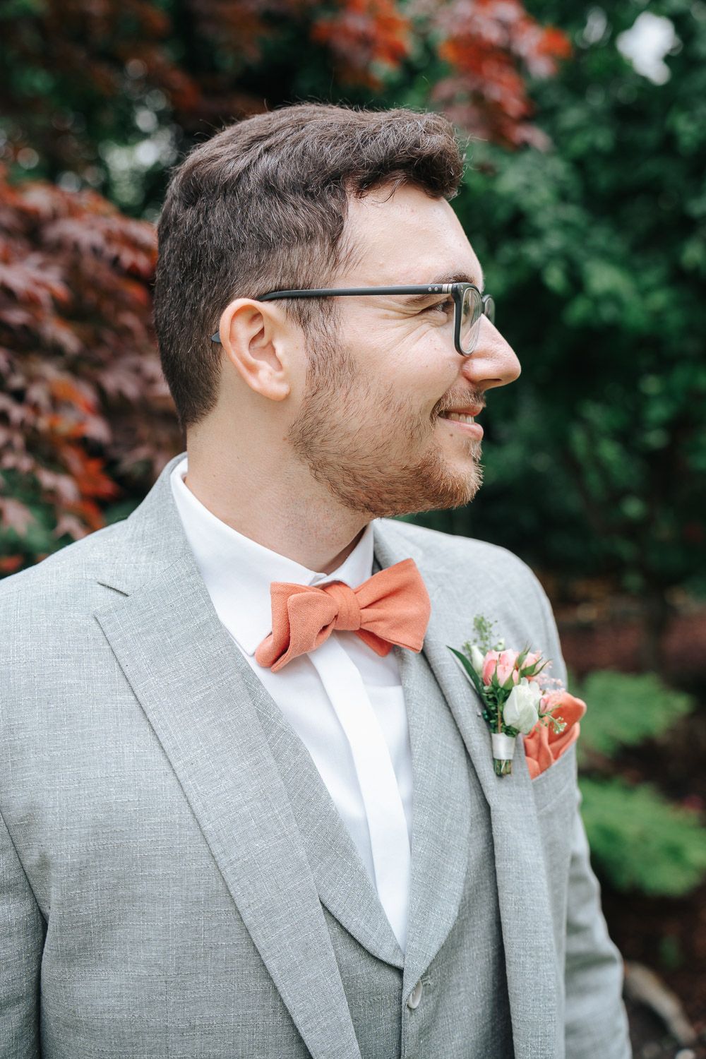 A smiling person in a light gray wedding suit with coral bow tie and boutonniere stands by autumn foliage.