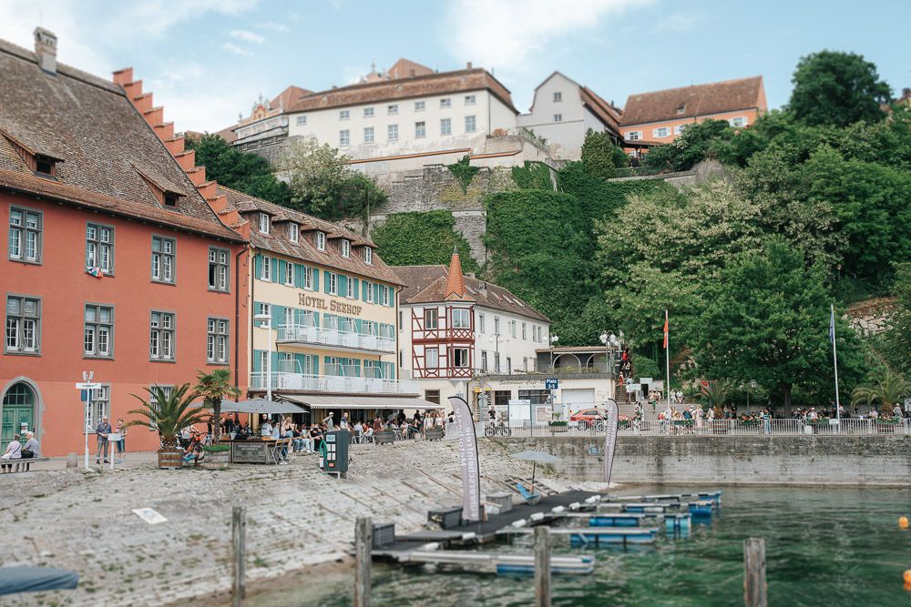 Colorful historic buildings and small boats line the waterfront in Meersburg, Germany with a hilltop castle in the background.