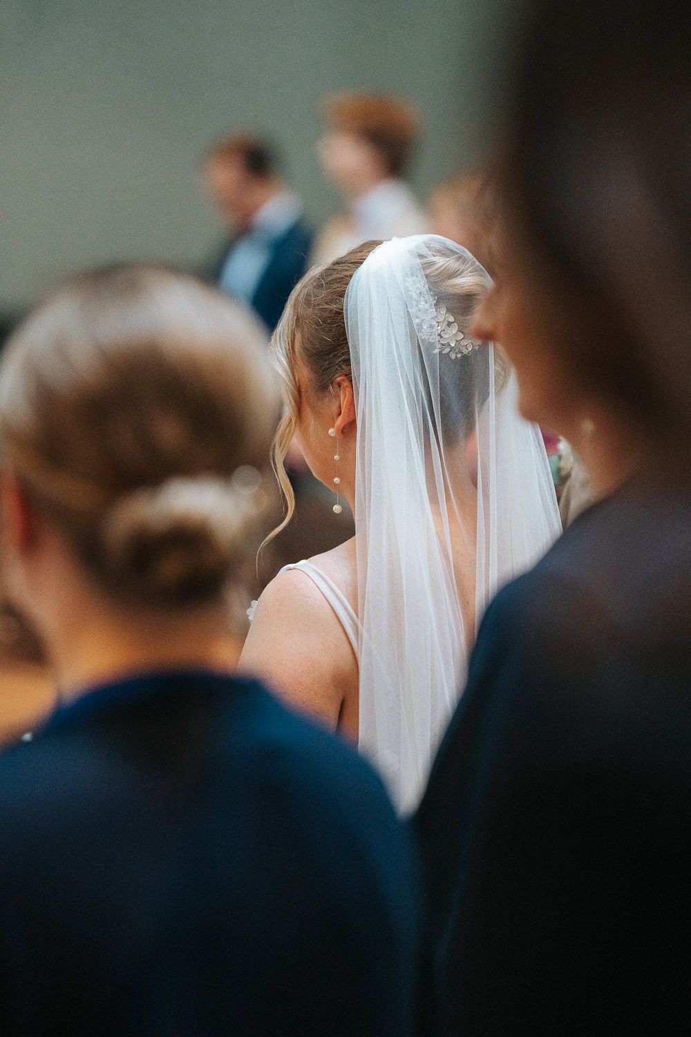 A white wedding veil flows elegantly from a bride's upswept hairstyle during a wedding ceremony.