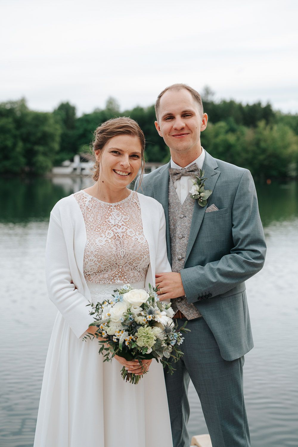 A couple poses by a lake in wedding attire with the bride wearing a lace gown and white cardigan holding a floral bouquet.